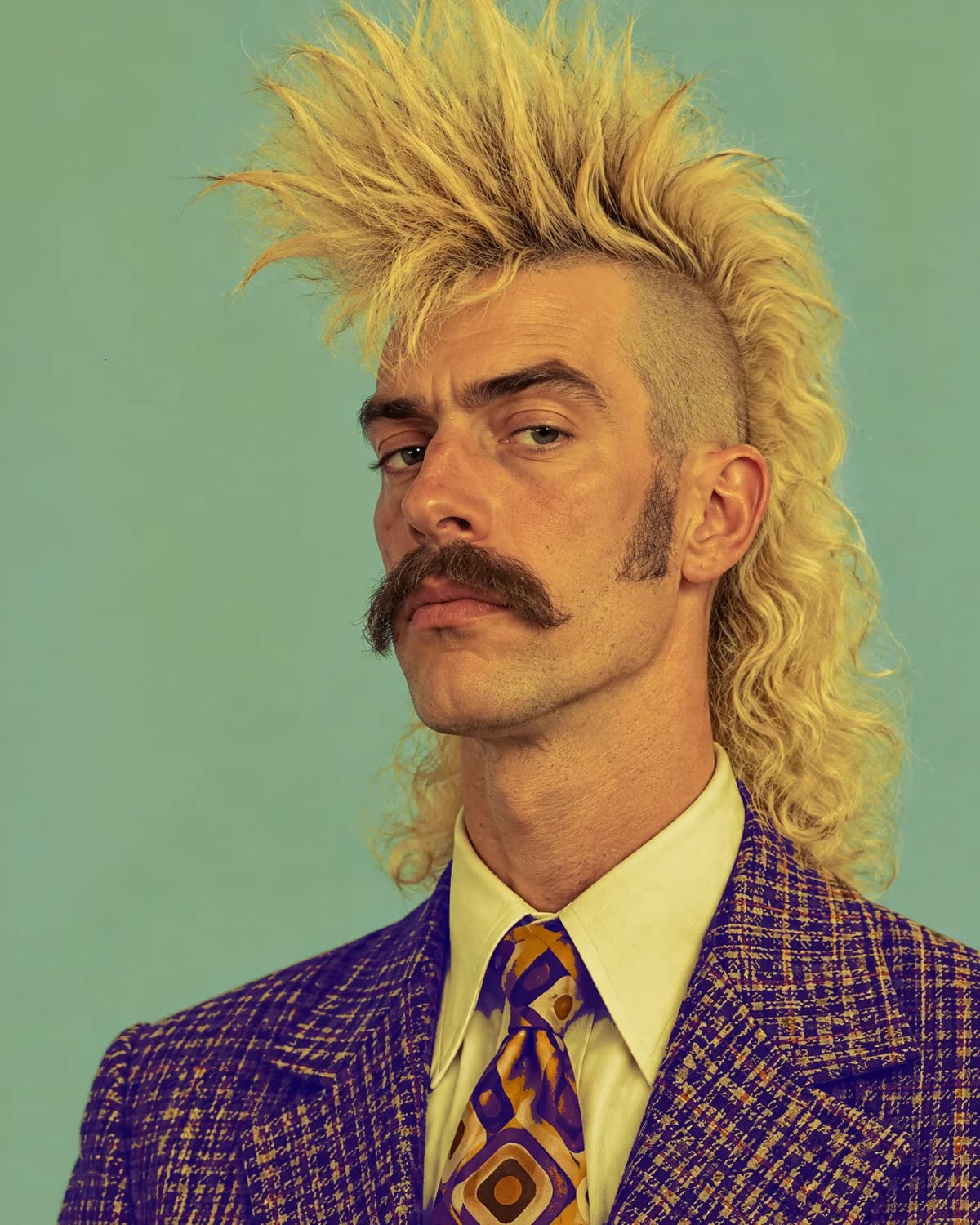 CB20, the host: blond spiky mullet, moustache, purple houndstooth suit, mustard tie